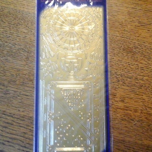 May include: A gold-colored metal stencil with a design of a dartboard and a trophy. The stencil is packaged in a clear plastic sleeve with a blue border.
