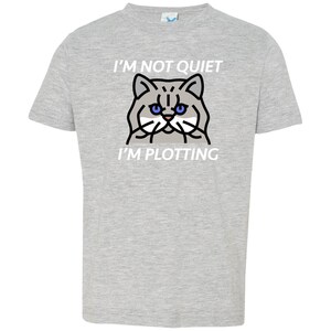 Pode incluir: Uma camiseta cinza com um gr&aacute;fico branco de um gato com olhos azuis e o texto "I'm not quiet I'm plotting".