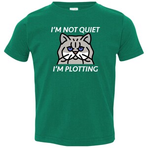 Pode incluir: Uma camiseta verde para crian&ccedil;as pequenas com uma ilustra&ccedil;&atilde;o de um gato branco e o texto "N&atilde;o estou quieto, estou tramando".
