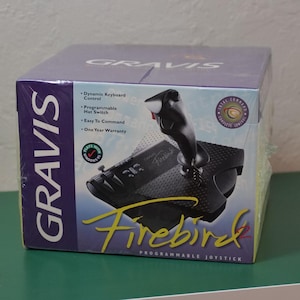 May include: A Gravis Firebird programmable joystick in its original purple and white cardboard packaging. The box features an image of the black joystick, the product name in yellow script, and text listing features like dynamic keyboard control.