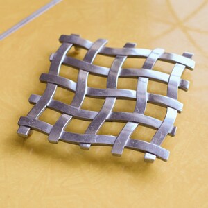 May include: A silver metal trivet with a woven design. The trivet is square shaped and has a shiny finish.