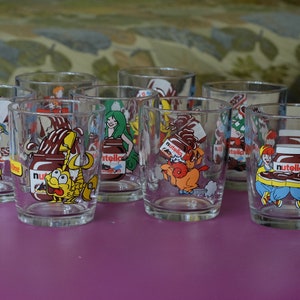 May include: Eight clear glass drinking glasses with colorful cartoon illustrations of people and animals enjoying Nutella hazelnut spread. The glasses feature the Nutella logo and the words "Nutella" and "Nuttiest" printed on them.