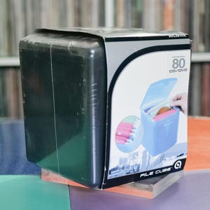 May include: A black plastic Atlantic File Cube box, designed to hold 80 CDs or DVDs. The box features a graphic of a blue file cube with discs inside. The box is on a multi-colored surface.
