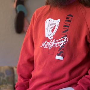 Może przedstawiać: Czerwona bluza z białym nadrukiem harfy i tekstem "GUINNESS" z podpisem "Arthur Guinness" pod spodem.