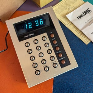 May include: A silver TimeTrac #2306 & #3300 appointment clock with a digital display showing 12:36. The clock has a variety of buttons including clear, cancel, date, timer, alarm, fur appt, and daily appt.