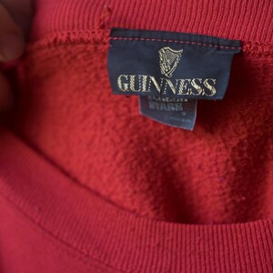 Może przedstawiać: Czerwona bluza z czarną metką z napisem "GUINNESS" złotymi literami.