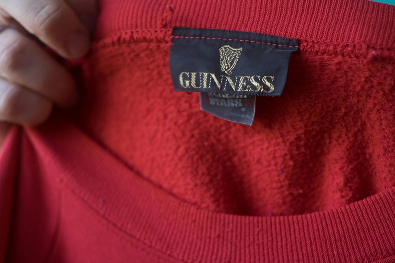 Może przedstawiać: Czerwona bluza z czarną metką z napisem "GUINNESS" złotymi literami.