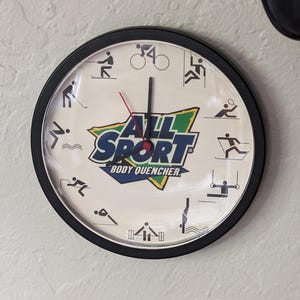 May include: A black and white clock with a white face and black hands. The clock has a green and white logo that says "All Sport Body Quencher". The clock face has various sports icons around the clock face.