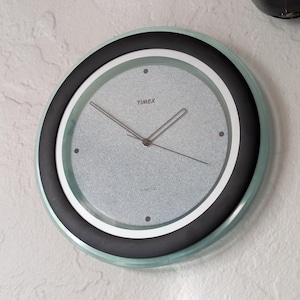 May include: A round, silver-faced wall clock with black hands and a black outer ring. The clock face has a textured, speckled appearance and the word "TIMEX" printed in black at the top.
