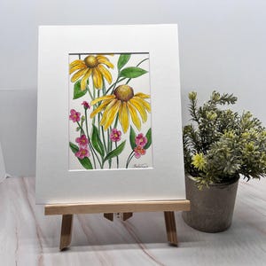 May include: A watercolor painting of yellow coneflowers and small pink flowers with green leaves. The artwork is displayed in a white mat and frame, resting on a small wooden easel. A small potted plant is to the right.