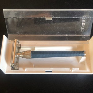 Puede incluir: Una caja de plástico blanca con una tapa de plástico transparente, que contiene una maquinilla de afeitar de seguridad plateada y azul con la palabra "Gillette" impresa en la tapa.