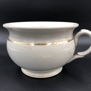 Peut inclure: Pot de chambre en céramique blanche avec une bordure dorée autour du haut. Le pot a une seule poignée et une base arrondie.