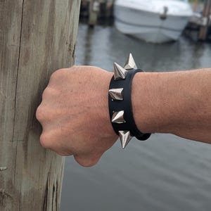 Może przedstawiać: Czarna skórzana bransoletka ze srebrnymi stożkowymi ćwiekami. Bransoletka jest noszona na nadgarstku, z drewnianym słupem w tle. W oddali widoczne są łodzie.