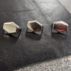 May include: Three silver-colored, hexagonal-shaped buttons are arranged in a row on a black leather surface. The buttons have a shiny, reflective finish, and are evenly spaced. The background is a textured, dark surface.