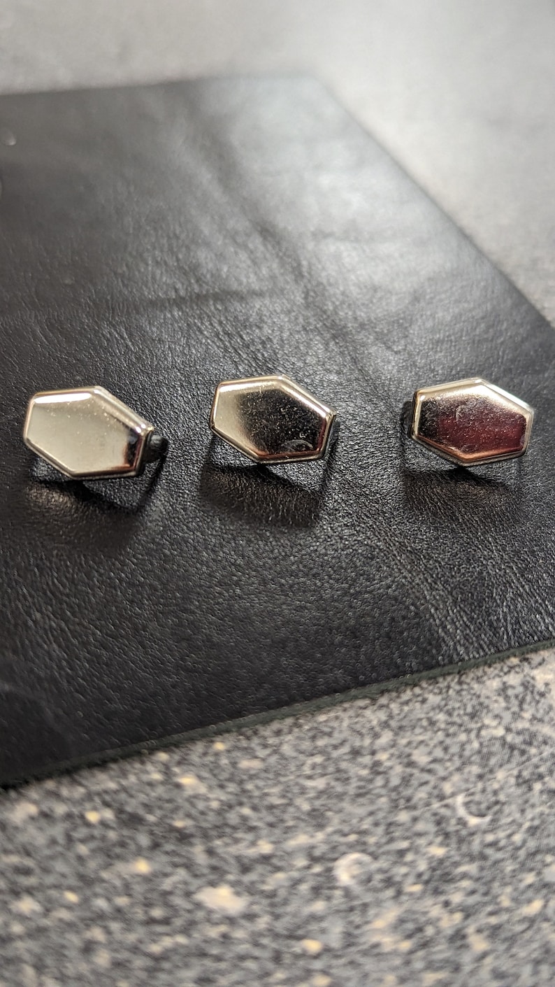 May include: Three silver-colored, hexagonal-shaped buttons are arranged in a row on a black leather surface. The buttons have a shiny, reflective finish, and are evenly spaced. The background is a textured, dark surface.
