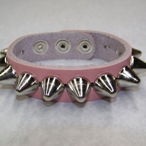 May include: A pink leather bracelet with silver metal spikes. The bracelet has three adjustable holes for sizing.
