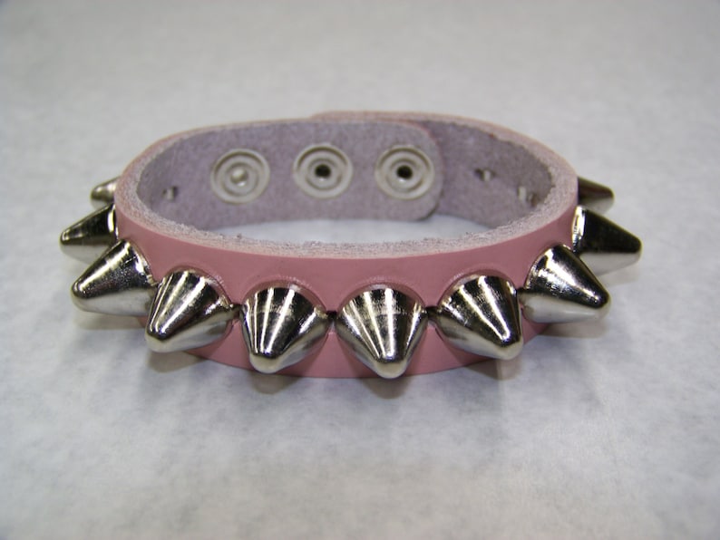 May include: A pink leather bracelet with silver metal spikes. The bracelet has three adjustable holes for sizing.