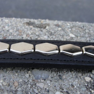 May include: A black leather bracelet with silver hexagonal studs. The bracelet is on a grey, pebbled surface.