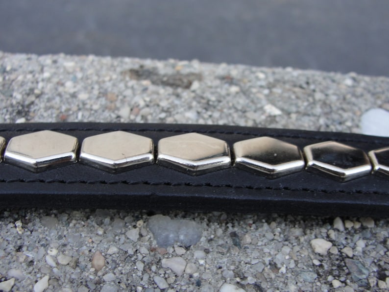 May include: A black leather bracelet with silver hexagonal studs. The bracelet is on a grey, pebbled surface.