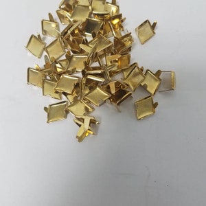 May include: A pile of small, gold-colored metal square-shaped fasteners with prongs. The fasteners are scattered on a white surface, and appear to be new.