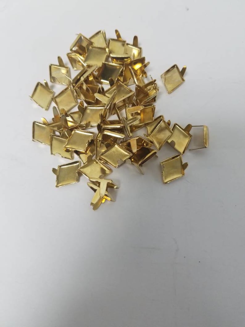 May include: A pile of small, gold-colored metal square-shaped fasteners with prongs. The fasteners are scattered on a white surface, and appear to be new.