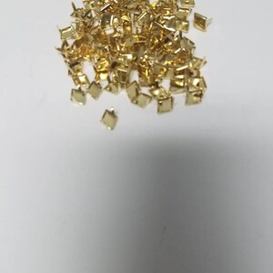 May include: A pile of small, gold-colored, square-shaped metal studs with short, pointed posts on a white background. The studs are likely used for decorative purposes or to attach materials.