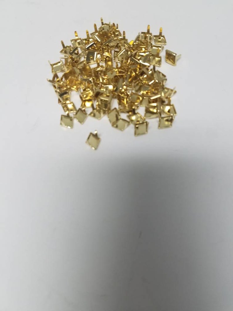 May include: A pile of small, gold-colored, square-shaped metal studs with short, pointed posts on a white background. The studs are likely used for decorative purposes or to attach materials.