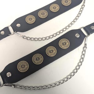 May include: Black leather accessory with silver chain and decorative shotgun shell accents. Each strap features three shell casings. The accessory is designed with a punk or alternative aesthetic.