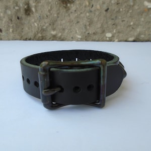 May include: A black leather bracelet with a silver buckle and a row of silver studs. The bracelet is on a white surface.
