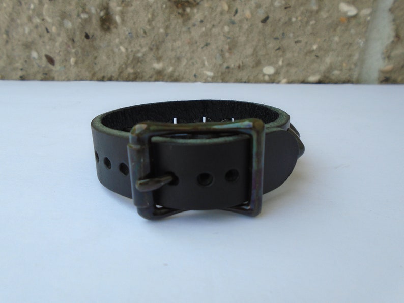 May include: A black leather bracelet with a silver buckle and a row of silver studs. The bracelet is on a white surface.