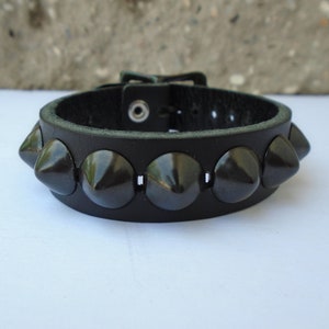 May include: A black leather bracelet with a buckle closure and a row of black pyramid-shaped studs.