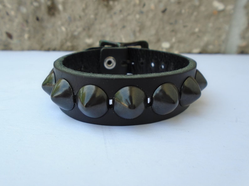 May include: A black leather bracelet with a buckle closure and a row of black pyramid-shaped studs.