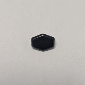May include: A small, black, hexagonal object with a glossy finish. The object is centered on a plain, light-colored background. The shape is a six-sided polygon, with slightly rounded edges.