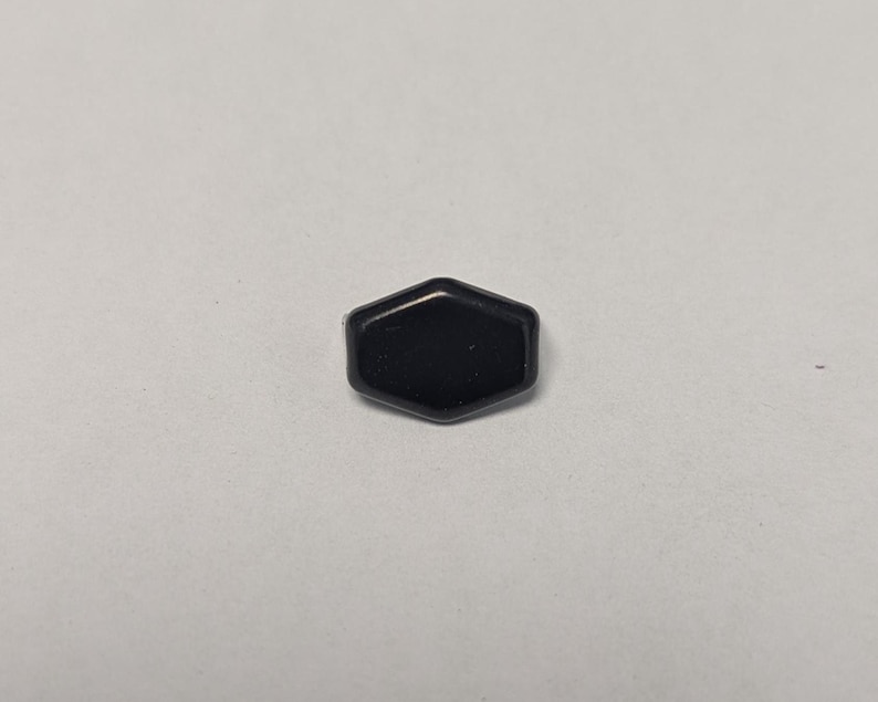 May include: A small, black, hexagonal object with a glossy finish. The object is centered on a plain, light-colored background. The shape is a six-sided polygon, with slightly rounded edges.
