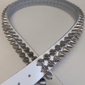 May include: White leather belt with two rows of silver cone-shaped studs and a row of rounded studs. The belt has a buckle and several holes for adjustment. A stylish accessory.