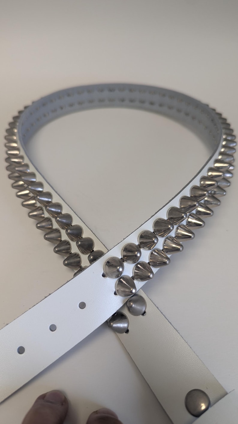 May include: White leather belt with two rows of silver cone-shaped studs and a row of rounded studs. The belt has a buckle and several holes for adjustment. A stylish accessory.