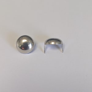 May include: Two small, shiny, silver-colored metal rivets. One is a complete, rounded dome shape. The other is a half-dome shape with two small, straight prongs extending downward. The rivets are on a white surface.