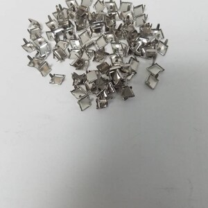 May include: A collection of small, silver-colored metal studs with square tops and short posts. The studs are scattered across a white surface, reflecting light. These decorative studs are likely used for crafting or embellishment.