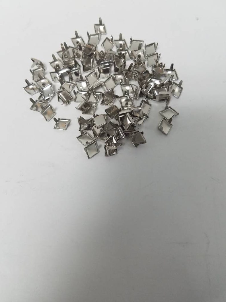 May include: A collection of small, silver-colored metal studs with square tops and short posts. The studs are scattered across a white surface, reflecting light. These decorative studs are likely used for crafting or embellishment.