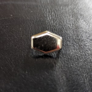 May include: A small, shiny, silver-colored, hexagonal object with a reflective surface. The object is centered on a dark, textured surface, possibly leather. The edges of the object are slightly rounded.