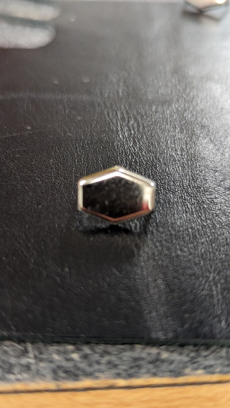 May include: A small, shiny, silver-colored, hexagonal object with a reflective surface. The object is centered on a dark, textured surface, possibly leather. The edges of the object are slightly rounded.
