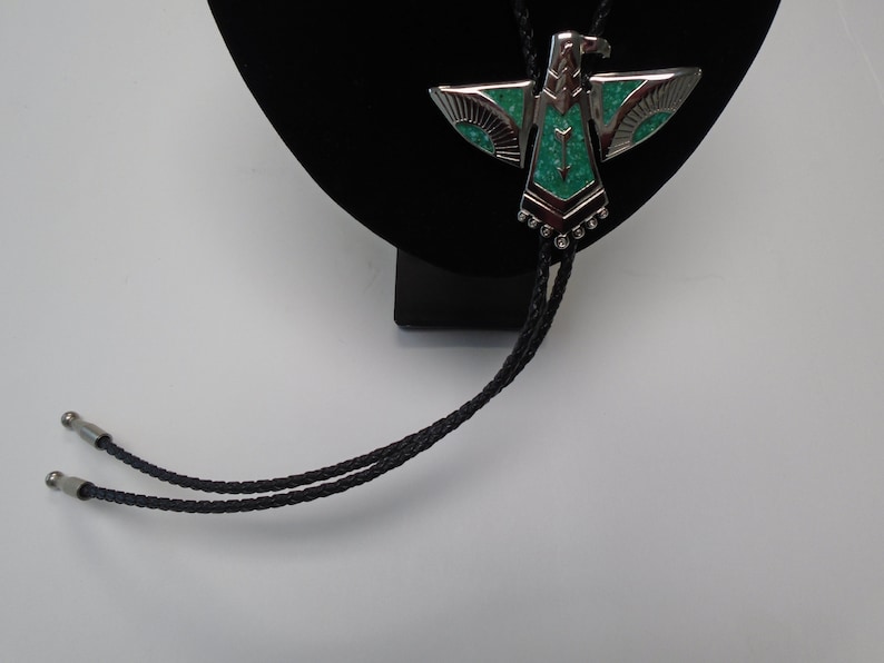 K&ouml;nnte beinhalten: Ein silberner Bolo-Schlips mit einem t&uuml;rkisfarbenen Adler-Design. Der Bolo-Schlips befindet sich auf einem schwarzen Samtst&auml;nder.