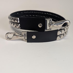 May include: Black leather strap with silver-colored pyramid studs and metal clasps. The strap has two layers of studs and a silver-colored clasp on each end. The strap is designed to be attached to a bag or other accessory.