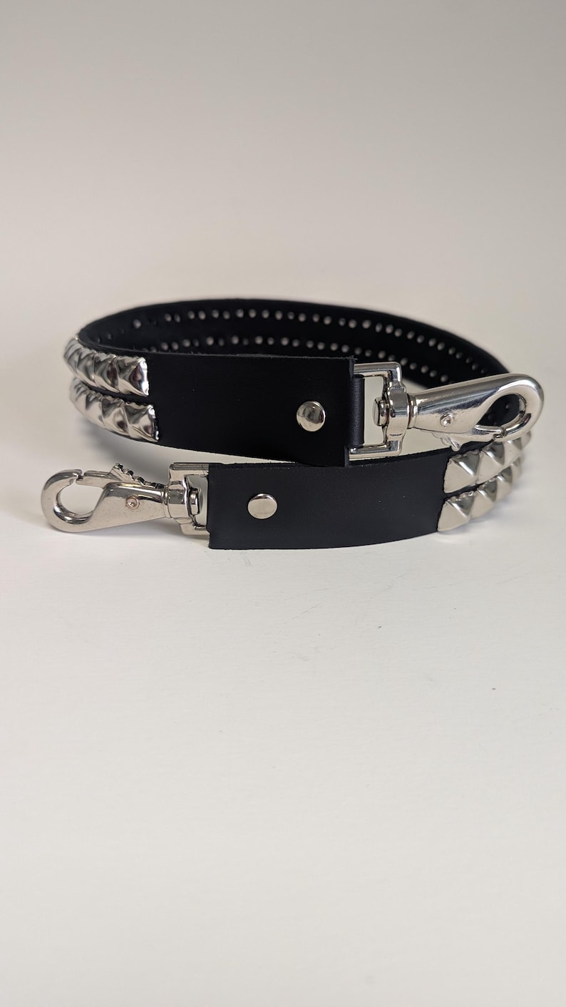 May include: Black leather strap with silver-colored pyramid studs and metal clasps. The strap has two layers of studs and a silver-colored clasp on each end. The strap is designed to be attached to a bag or other accessory.
