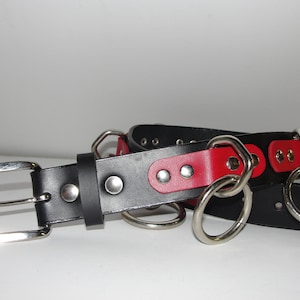 May include: A black leather belt with a red leather accent and multiple silver metal rings. The belt has a silver buckle.