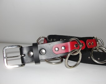 Puede incluir: Un cinturón de cuero negro con un acento de cuero rojo y múltiples anillos de metal plateado. El cinturón tiene una hebilla plateada.