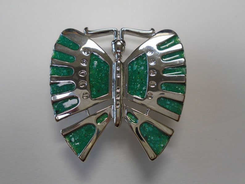 K&ouml;nnte beinhalten: Eine silberne Brosche in Form eines Schmetterlings mit gr&uuml;nem Glitzer-Emaille. Der Schmetterling hat ein detailliertes Design mit einem erhabenen K&ouml;rper und Fl&uuml;geln.