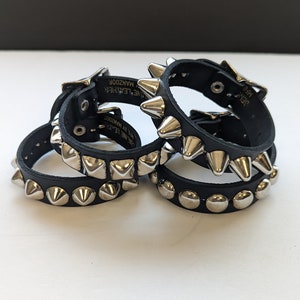 May include: Four black leather bracelets with silver pyramid-shaped studs. The bracelets have a buckle closure and the text "Genuine Leather" and "Manzoor" is printed on the back of one of the bracelets.