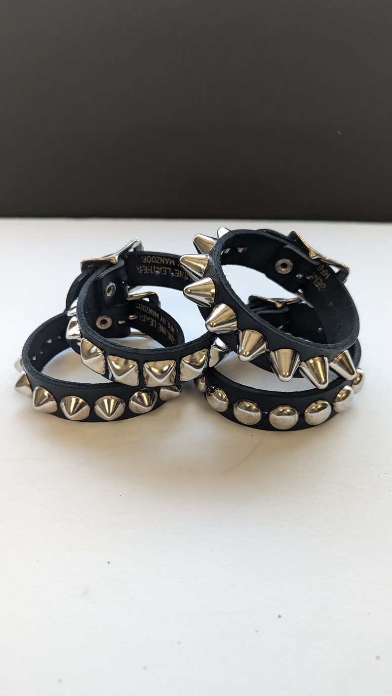 May include: Four black leather bracelets with silver pyramid-shaped studs. The bracelets have a buckle closure and the text "Genuine Leather" and "Manzoor" is printed on the back of one of the bracelets.