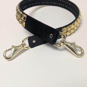 May include: Black handbag strap with gold-colored pyramid studs and gold-tone metal clasps. The strap is made of black leather and has a series of small holes along the top edge. The clasps are attached to the strap with metal rivets.